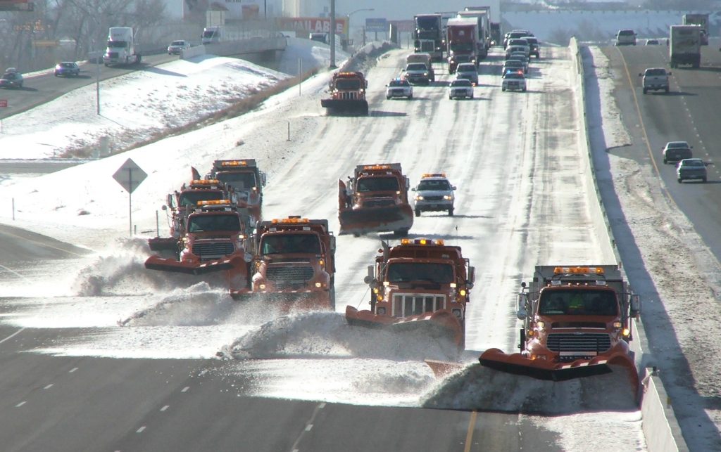 7 snow plows travel in tandem on freeway lanes pushing snow.