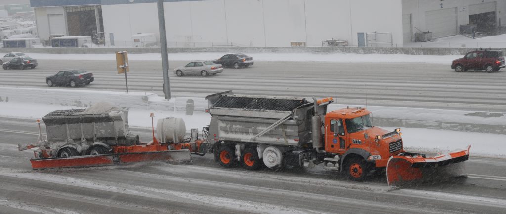Profile view of a large snow plow with the tow plow attachment on a snowy freeway.