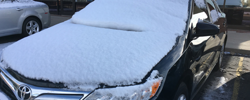 Snow covers the hood and windshield of a parked car.