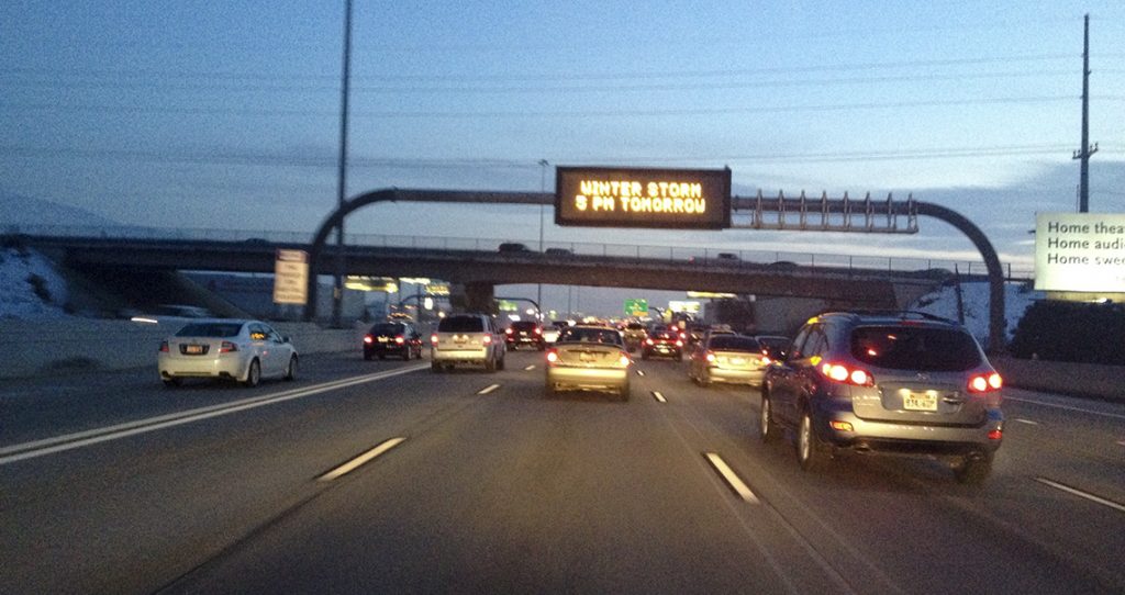 Vehicles drive on a freeway while the overhead sign says "Winter Storm Tomorrow 5 pm"