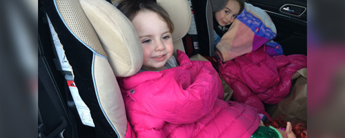 Two young girls sit in car seats with jackets placed over them.