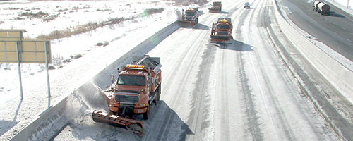 Four snow plows are on a sunny snowy road pushing snow.