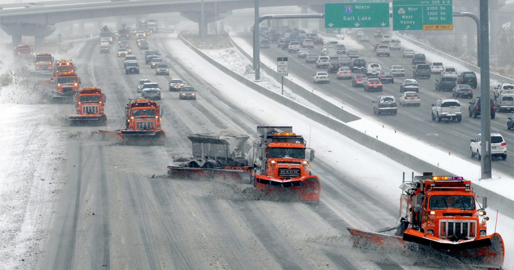 eight snow plows travel in tandem pushing snow off a snowy road.