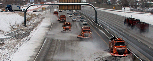 Five snow plows are lined up on a freeway pushing snow.